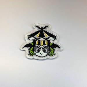 May include: An embroidered patch featuring a character with wild green hair, pale skin, and dark eyes, wearing a black and white striped top hat with yellow accents and a small black bat silhouette on top, all set against a white glittery border.