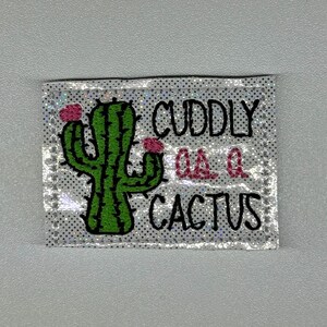 May include: A rectangular patch with a green cactus illustration and the words "CUDDLY QA Q CACTUS" in black and pink. The patch has a silver, glittery background with small black dots.