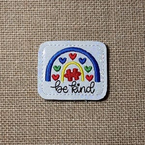 May include: A square patch with rounded corners featuring a white background with small blue dots. The design includes a blue and yellow rainbow, small red, blue, and green hearts, a red puzzle piece, and the black cursive text be kind.