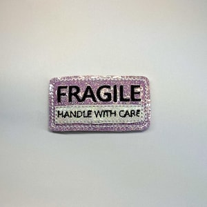 May include: A rectangular patch with a lavender glitter finish featuring the black embroidered text FRAGILE in the center and the text HANDLE WITH CARE embroidered in black on a white glitter strip positioned below the main word.