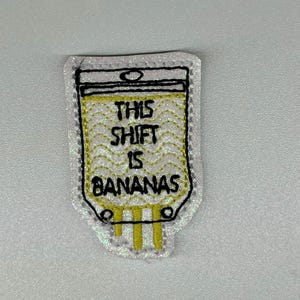 May include: A white and glittery patch with a black outline and the text "THIS SHIFT IS BANANAS" in black. The inside of the patch is yellow with a wavy pattern. The patch has three yellow lines at the bottom.