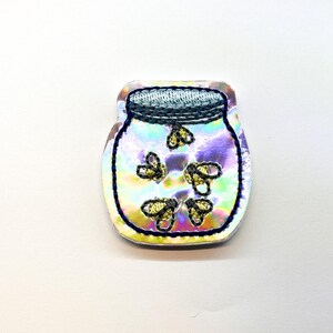 May include: A holographic jar-shaped patch featuring an embroidered light blue lid and a dark blue stitched outline. Inside the jar, five small yellow embroidered fireflies are depicted against the iridescent, multi-colored surface.