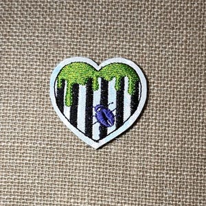 May include: A heart-shaped embroidered patch featuring a black and white vertical striped pattern. The top of the heart is covered in a bright green dripping liquid design, and a small purple beetle is centered on the lower half of the heart.