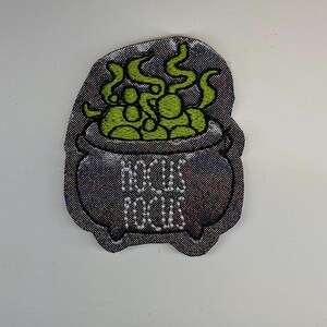 May include: A black embroidered patch shaped like a cauldron with the text HOCUS POCUS stitched in white thread. The cauldron contains a bubbling green liquid with swirling steam rising from the top, all rendered in detailed embroidery thread.