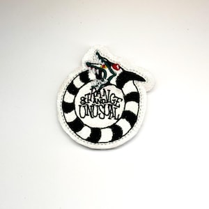 May include: A circular embroidered patch featuring a black and white striped snake coiled into a ring. The snake has a red eye and green accents. The text "STRANGE AND UNUSUAL" is stitched in black thread in the center of the coiled snake design.