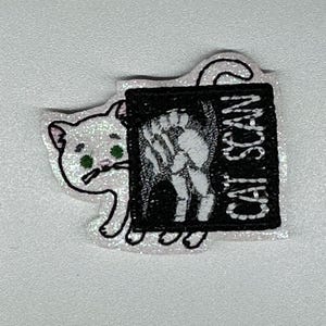 May include: A glittery white cat-shaped patch with green eyes peeking out from behind a black rectangle. The rectangle features a cat skeleton image and the words "CAT SCAN" in white.