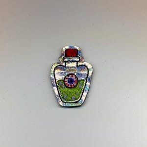 May include: An embroidered patch shaped like a potion bottle with a holographic silver finish. The bottle contains green liquid with small purple dots and a central red and blue eyeball graphic. It features a red stopper at the top.