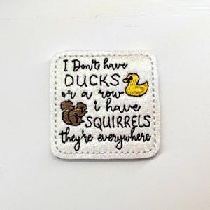 May include: A square white patch with rounded corners featuring black embroidered text that reads I Don't have DUCKS or a row i have SQUIRRELS they're everywhere. The patch includes a small yellow embroidered duck and a brown embroidered squirrel.