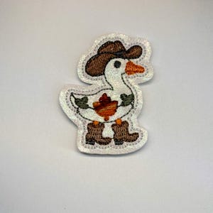 May include: A white embroidered patch featuring a goose wearing a brown cowboy hat and brown cowboy boots. The goose has an orange beak and a small orange maple leaf design on its chest, all set against a shimmering white glittery background.