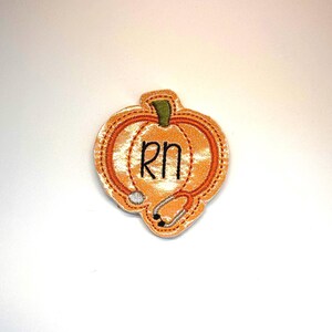 May include: An orange pumpkin-shaped embroidered patch featuring a green stem and a stethoscope design stitched in orange thread. The letters RN are embroidered in black in the center of the pumpkin, with a silver-colored stethoscope head.