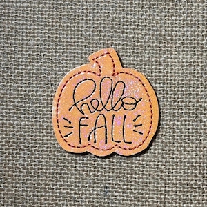 May include: A pumpkin-shaped patch with a glittery orange finish and dark red stitched borders. The text "hello FALL" is embroidered in black thread across the center of the pumpkin, featuring a stem at the top and decorative stitched lines.