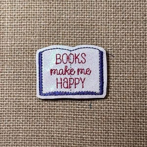 May include: A white, glittery, book-shaped embroidered patch with a dark blue border. The text "BOOKS make me HAPPY" is stitched in red thread across the center of the patch, which is shaped like an open book with a visible spine line.