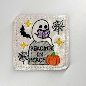 May include: A square, glittery patch with a Halloween theme. It features a white ghost reading a purple book atop a tombstone that reads "READING IN PEACE." Other elements include a bat, spiderwebs, stars, and a pumpkin.