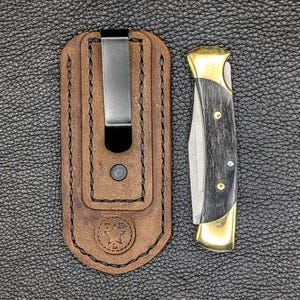 K&ouml;nnte beinhalten: Eine braune Leder-Messerscheide mit einem Metallclip neben einem Klappmesser. Das Messer hat einen schwarzen Griff mit Messingakzenten und eine silberne Klinge. Die Scheide hat ein Sternlogo.