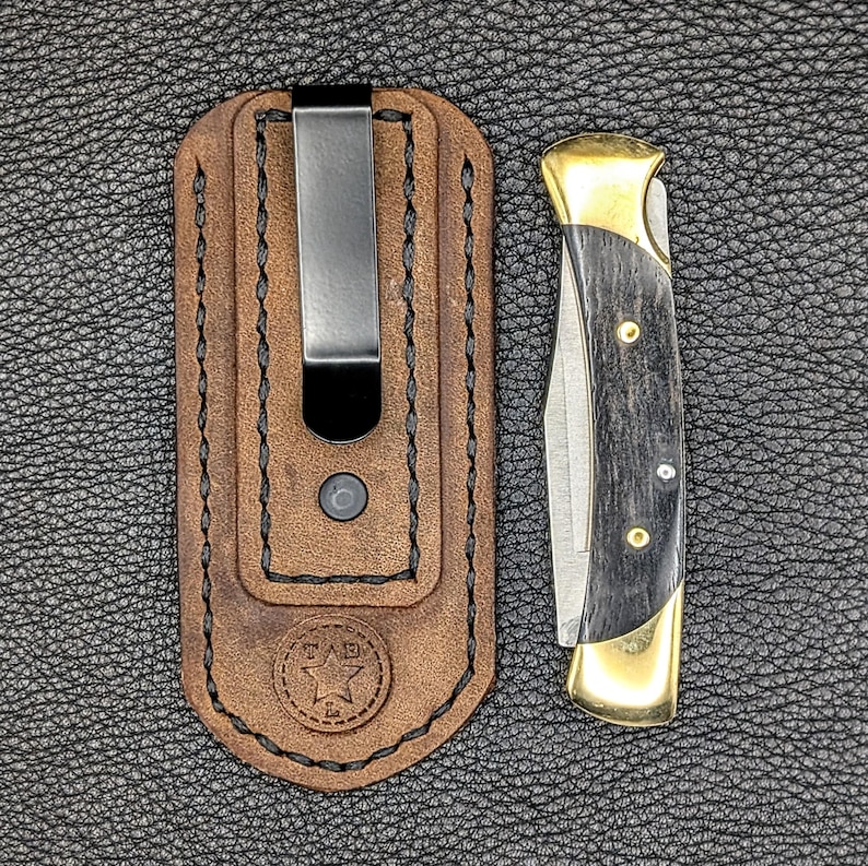 K&ouml;nnte beinhalten: Eine braune Leder-Messerscheide mit einem Metallclip neben einem Klappmesser. Das Messer hat einen schwarzen Griff mit Messingakzenten und eine silberne Klinge. Die Scheide hat ein Sternlogo.