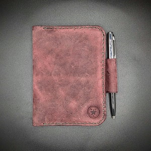 May include: A burgundy leather notebook with a pen holder and a star logo. The notebook has a stitched edge and a distressed look.