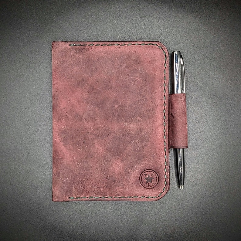 May include: A burgundy leather notebook with a pen holder and a star logo. The notebook has a stitched edge and a distressed look.