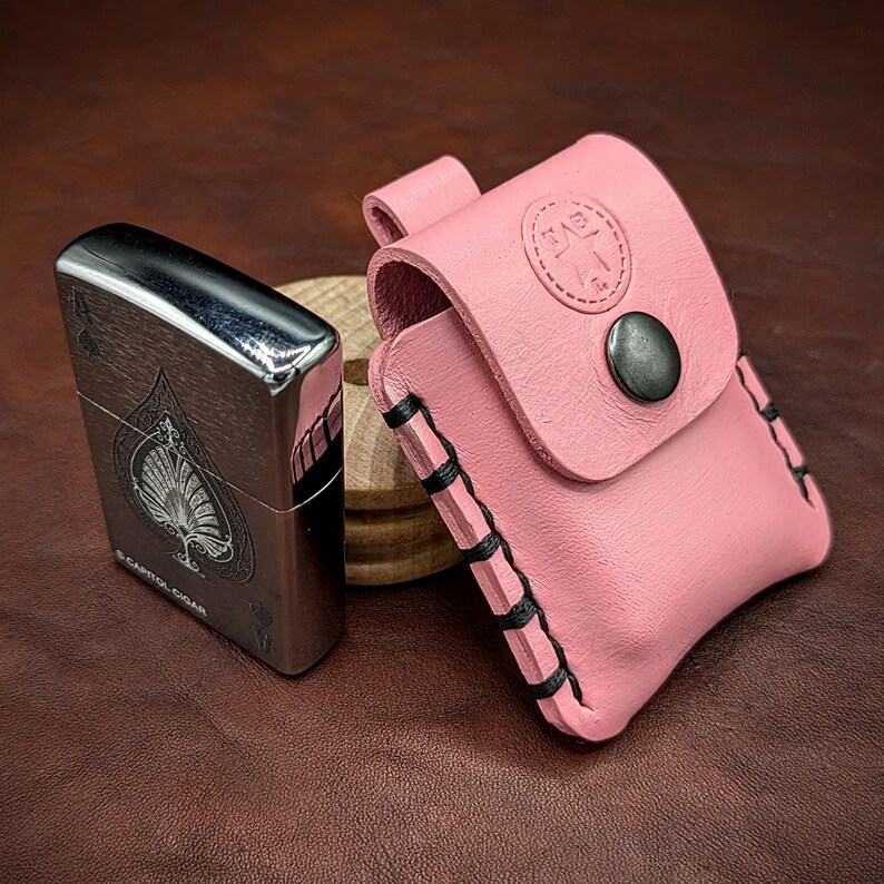 May include: A pink leather lighter case with black stitching and a silver button. The case has a small embossed logo on the front. A silver lighter with an ace of spades design is next to the case.