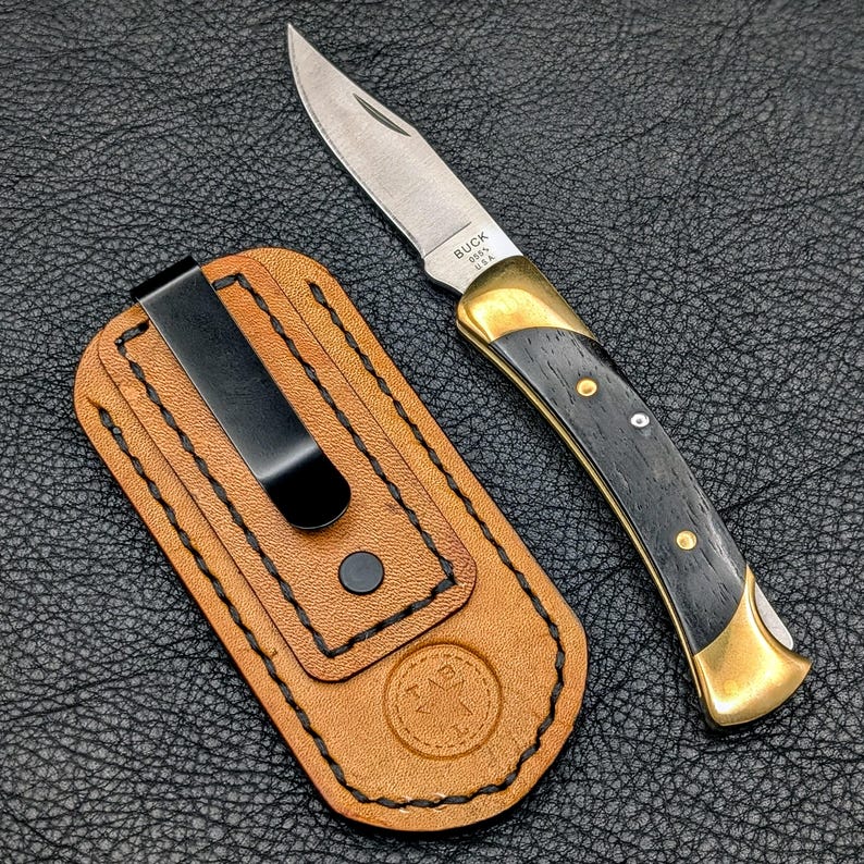 K&ouml;nnte beinhalten: Ein Buck-Klappmesser mit silberner Klinge und einem schwarz-goldenen Griff, neben einer braunen Lederscheide mit schwarzem Clip. Das Messer hat den Text "BUCK 0605 USA" auf der Klinge.