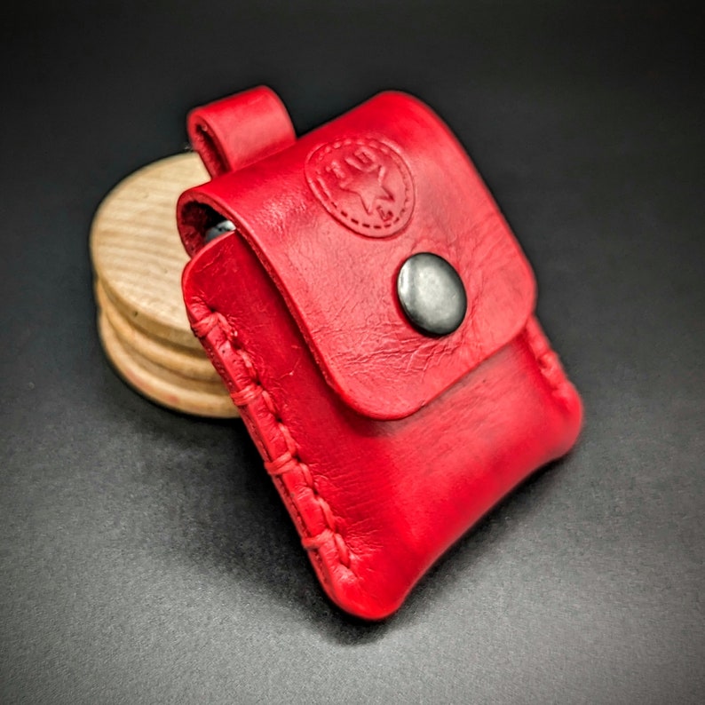 May include: A red leather case with a snap closure and a stitched design. The case has a small metal button on the front and a logo embossed on the top.