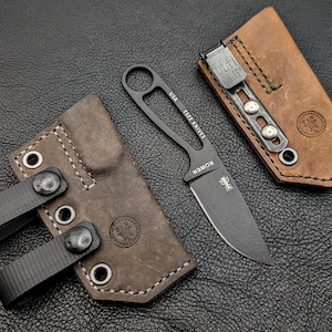 May include: A black fixed-blade knife with a leather sheath and a brown leather sheath with a metal clip. The knife has the text "USA ESEE KNIVES" and "ROWEN" on the blade. The sheaths are brown with black stitching.
