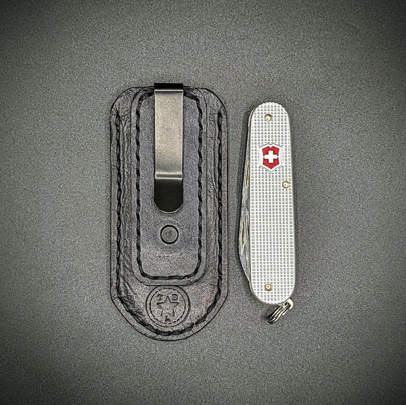 K&ouml;nnte beinhalten: Eine schwarze Leder-Taschenmesserscheide mit einem Metallclip neben einem silbernen Taschenmesser mit einem rot-wei&szlig;en Schweizer Kreuz-Emblem. Das Taschenmesser hat ein strukturiertes, gitterartiges Muster auf dem Griff.