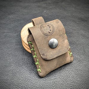 May include: A small, brown leather pouch with green stitching. The pouch has a flap with a snap closure and a metal button. The pouch is embossed with the text "TID" and a star.