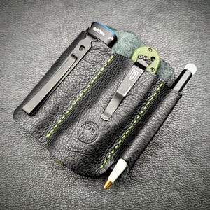 May include: Black leather EDC organizer with green stitching, holding a pen, knife, and flashlight. The organizer has a metal clip and a stamped emblem. The flashlight has the text "Arkflex".