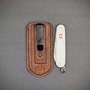 K&ouml;nnte beinhalten: Eine braune Leder-Taschenmesser-Scheide mit einem schwarzen Metallclip neben einem silbernen und wei&szlig;en Taschenmesser mit einem roten Kreuzemblem. Die Scheide hat eine gen&auml;hte Bord&uuml;re und ein gestempeltes Logo.