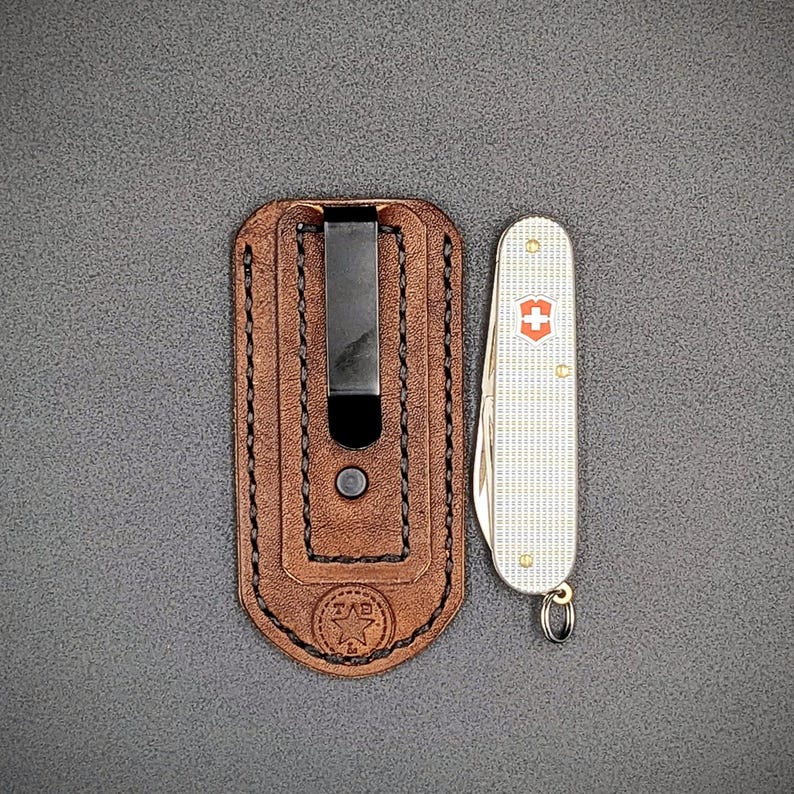 K&ouml;nnte beinhalten: Eine braune Leder-Taschenmesser-Scheide mit einem schwarzen Metallclip neben einem silbernen und wei&szlig;en Taschenmesser mit einem roten Kreuzemblem. Die Scheide hat eine gen&auml;hte Bord&uuml;re und ein gestempeltes Logo.