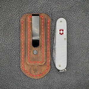 K&ouml;nnte beinhalten: Eine braune Leder-Taschenmesser-Scheide mit roten N&auml;hten und einem Metallclip, neben einem silbernen Schweizer Taschenmesser mit rotem und wei&szlig;em Emblem. Die Artikel befinden sich auf einer strukturierten schwarzen Oberfl&auml;che.