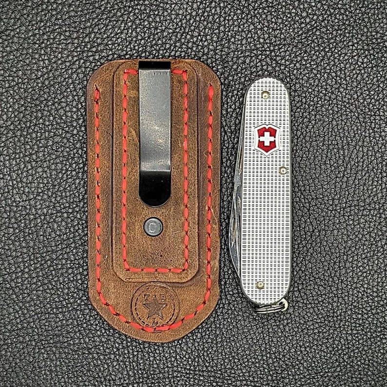 K&ouml;nnte beinhalten: Eine braune Leder-Taschenmesser-Scheide mit roten N&auml;hten und einem Metallclip, neben einem silbernen Schweizer Taschenmesser mit rotem und wei&szlig;em Emblem. Die Artikel befinden sich auf einer strukturierten schwarzen Oberfl&auml;che.