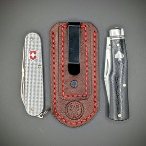 K&ouml;nnte beinhalten: Eine Auswahl an Taschenmessern und eine Lederscheide. Die Messer umfassen ein silbernes Schweizer Taschenmesser mit rotem Kreuz, ein schwarz-silbernes Messer mit Pik-Symbol und eine braune Lederscheide mit roten N&auml;hten und Metallclip.