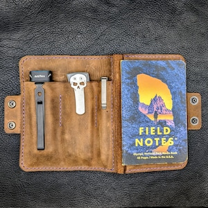 May include: Open brown leather notebook with a Field Notes memo book and three metal tools. The Field Notes book has a blue cover with a landscape scene. The leather case has purple stitching and snap closures.
