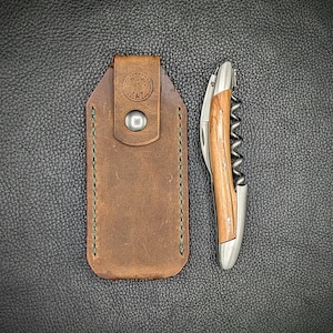 May include: A brown leather case with a stitched edge and a metal button. A silver corkscrew with a wooden handle is next to the case.