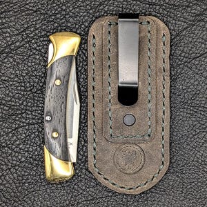 K&ouml;nnte beinhalten: Ein Klappmesser mit goldfarbener Zwinge und einem dunklen Holzmaserungsgriff. Das Messer liegt neben einer braunen Lederscheide mit einem Metallclip. Die Scheide hat gr&uuml;ne N&auml;hte und ein gepr&auml;gtes Emblem.