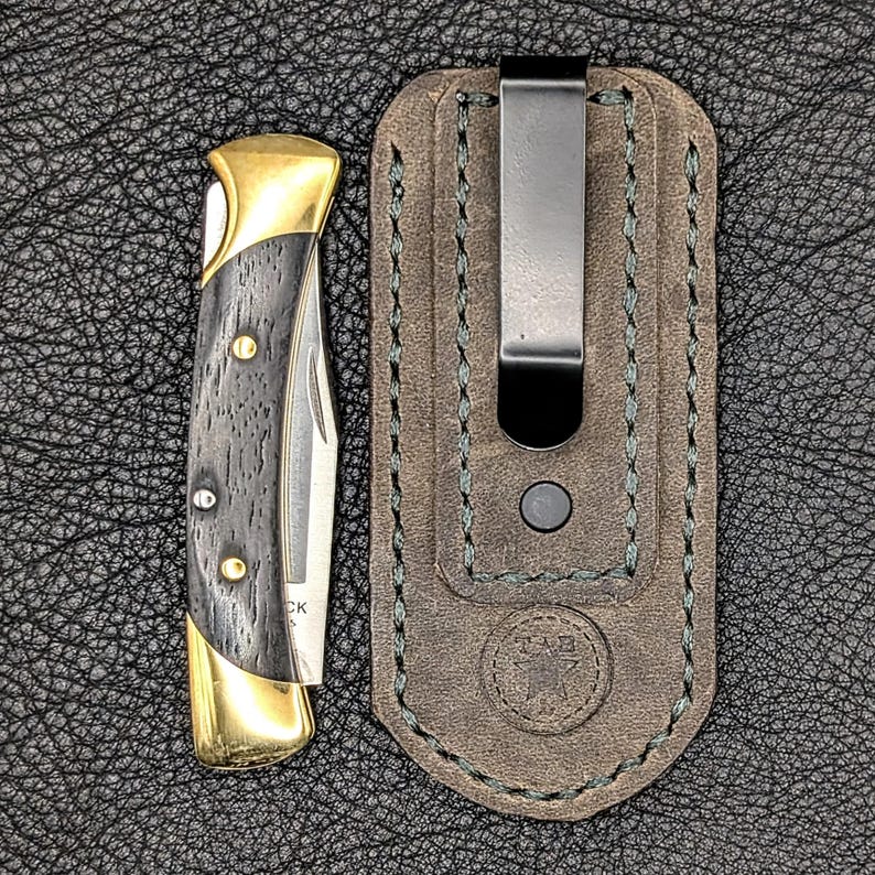 K&ouml;nnte beinhalten: Ein Klappmesser mit goldfarbener Zwinge und einem dunklen Holzmaserungsgriff. Das Messer liegt neben einer braunen Lederscheide mit einem Metallclip. Die Scheide hat gr&uuml;ne N&auml;hte und ein gepr&auml;gtes Emblem.