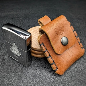 May include: A brown leather lighter case with a stitched edge and a silver button. The case is sitting on a wooden coaster next to a silver lighter with a black ace of spades design and the text "Capitol Cigar".