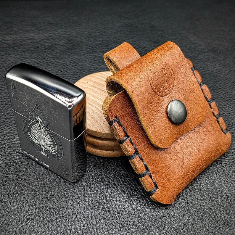 May include: A brown leather lighter case with a stitched edge and a silver button. The case is sitting on a wooden coaster next to a silver lighter with a black ace of spades design and the text "Capitol Cigar".