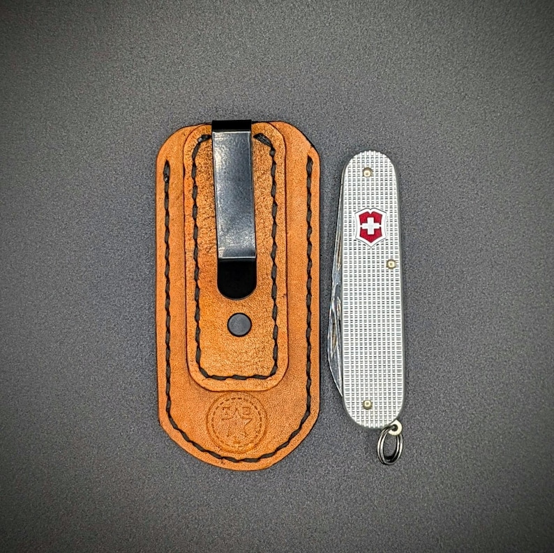 K&ouml;nnte beinhalten: Eine hellbraune Leder-Taschenmesser-Scheide mit einem schwarzen Metallclip neben einem silbernen Taschenmesser mit einem roten Kreuz-Emblem. Die Scheide hat ein gepr&auml;gtes Logo und ist mit schwarzem Faden gen&auml;ht.