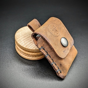 May include: A brown leather pouch with a stitched design and a metal snap closure. The pouch is sitting on a wooden surface.