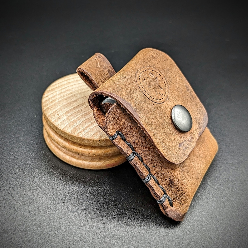 May include: A brown leather pouch with a stitched design and a metal snap closure. The pouch is sitting on a wooden surface.