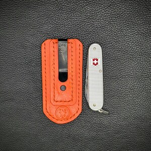 K&ouml;nnte beinhalten: Eine orangefarbene Leder-Taschenmesserh&uuml;lle mit einem schwarzen Clip neben einem silbernen Schweizer Taschenmesser mit einem roten Kreuzemblem. Die H&uuml;lle hat ein Sternemblem und den Text "ZAB" gepr&auml;gt.