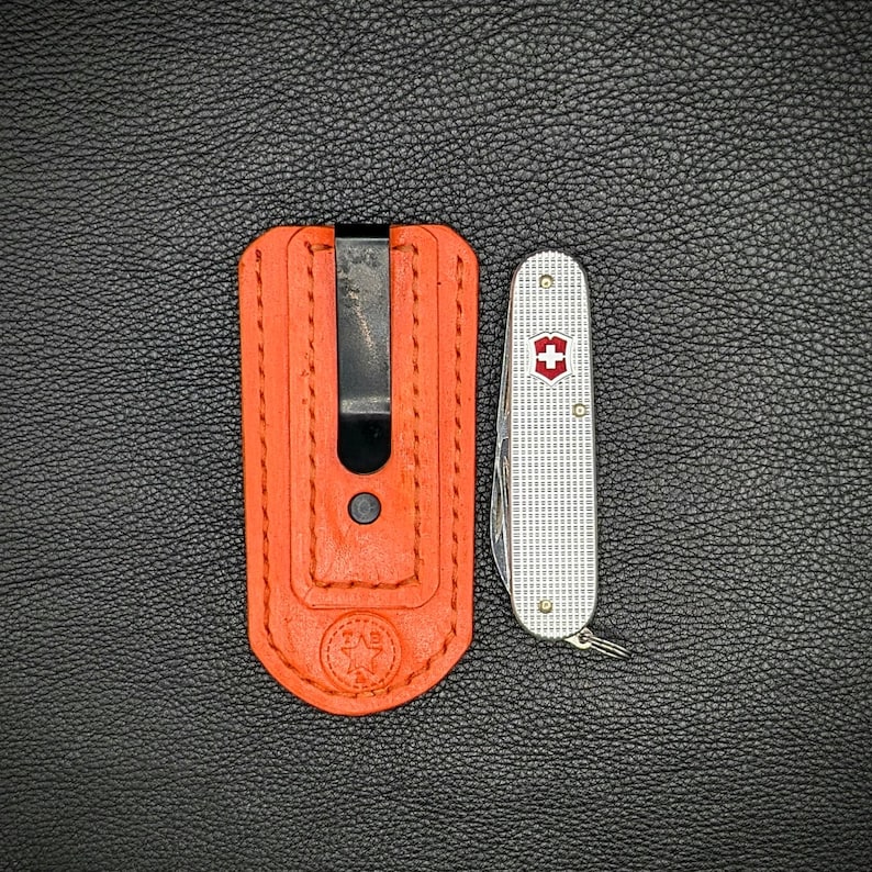 K&ouml;nnte beinhalten: Eine orangefarbene Leder-Taschenmesserh&uuml;lle mit einem schwarzen Clip neben einem silbernen Schweizer Taschenmesser mit einem roten Kreuzemblem. Die H&uuml;lle hat ein Sternemblem und den Text "ZAB" gepr&auml;gt.