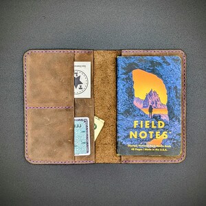May include: A brown leather notebook cover with a purple stitch detail. The cover has three slots for cards or cash. A Field Notes notebook is inside the cover. The cover is open and the notebook is visible. The notebook cover is made of leather and has a rustic look. The notebook is a Field Notes brand notebook with a design of a mountain and a cave on the front. The text on the notebook reads "FIELD NOTES" and "Olympic National Park Memo Book". The text also reads "48 Pages / Made in the U.S.A."