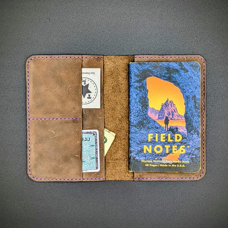 May include: A brown leather notebook cover with a purple stitch detail. The cover has three slots for cards or cash. A Field Notes notebook is inside the cover. The cover is open and the notebook is visible. The notebook cover is made of leather and has a rustic look. The notebook is a Field Notes brand notebook with a design of a mountain and a cave on the front. The text on the notebook reads "FIELD NOTES" and "Olympic National Park Memo Book". The text also reads "48 Pages / Made in the U.S.A."