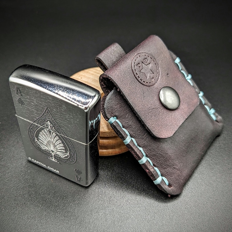May include: A silver lighter with a black ace of spades design and the text "Capitol Cigar". The lighter is next to a brown leather case with blue stitching and a star design.