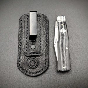 K&ouml;nnte beinhalten: Eine schwarze Leder-Taschenmesser-Scheide mit einem Metallclip neben einem geschlossenen Taschenmesser. Das Messer hat einen schwarz-silbernen Griff mit einem Pik-Symbol. Die Scheide hat ein Stern-Emblem.