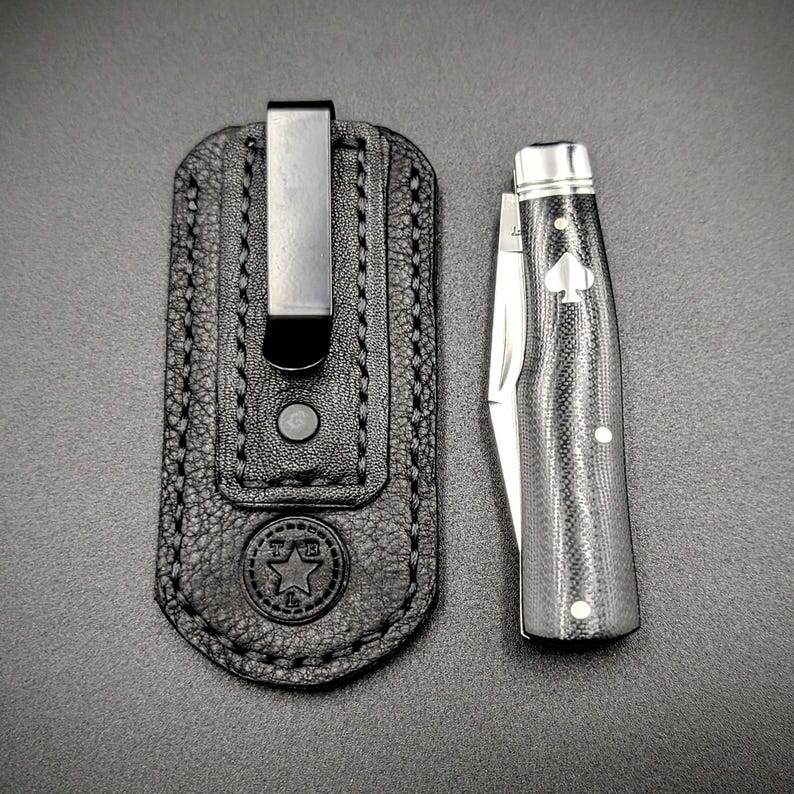 K&ouml;nnte beinhalten: Eine schwarze Leder-Taschenmesser-Scheide mit einem Metallclip neben einem geschlossenen Taschenmesser. Das Messer hat einen schwarz-silbernen Griff mit einem Pik-Symbol. Die Scheide hat ein Stern-Emblem.