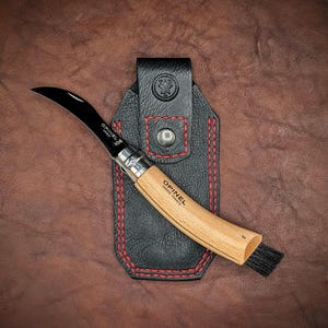 May include: A black leather sheath with red stitching holds a curved Opinel knife with a wooden handle and a small brush. The knife has a black blade with the text "Opinel  Made in France" on it.